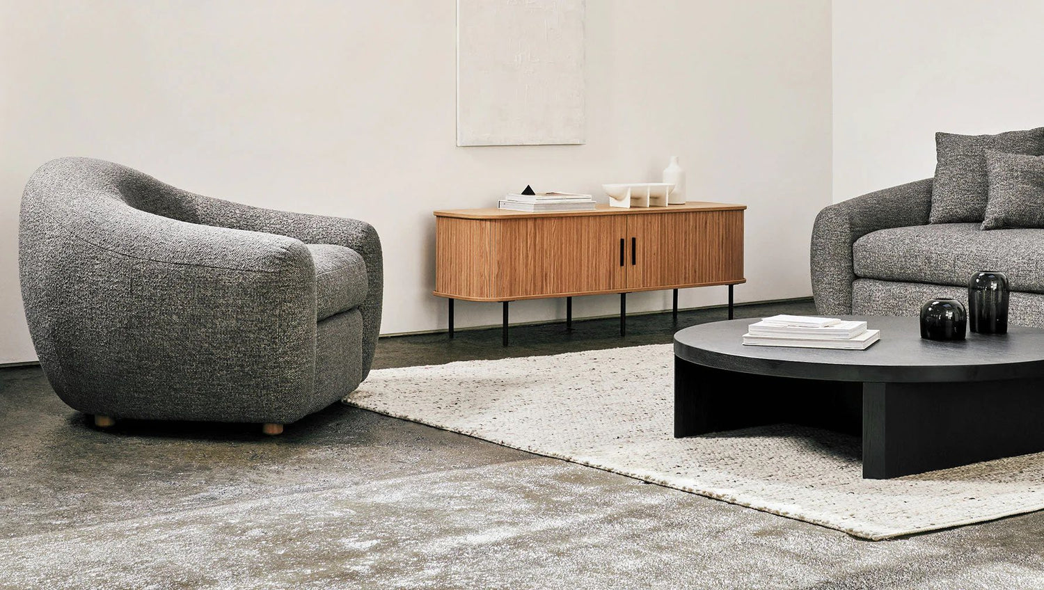 A modern living room featuring a gray upholstered armchair and sofa, a rectangular wooden cabinet with angled legs, and a low, round black coffee table on a light-colored rug. The walls are white and there's minimal decor, creating a clean and minimalist aesthetic.