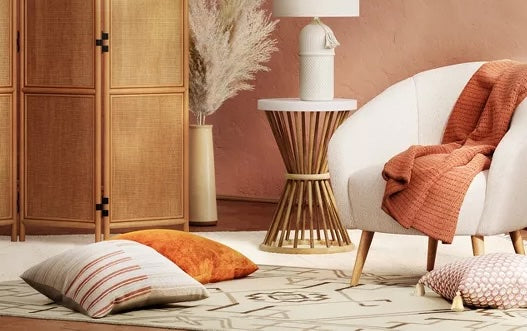 A cozy living room with a white armchair draped with an orange blanket. A modern wooden side table holds a decorative vase and lamp. Cushions in various colors and patterns are on the light-colored patterned rug. A wicker room divider and pampas grass are in the background.