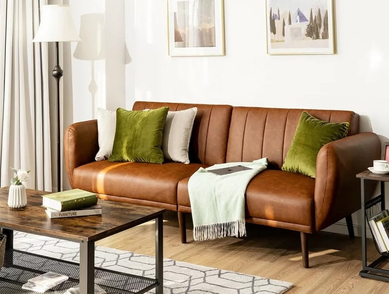 A cozy living room featuring a brown leather sofa adorned with green and beige cushions, and a light green throw blanket. A rustic wooden coffee table sits in front, and a floor lamp and framed artworks decorate the background. Natural light fills the room.