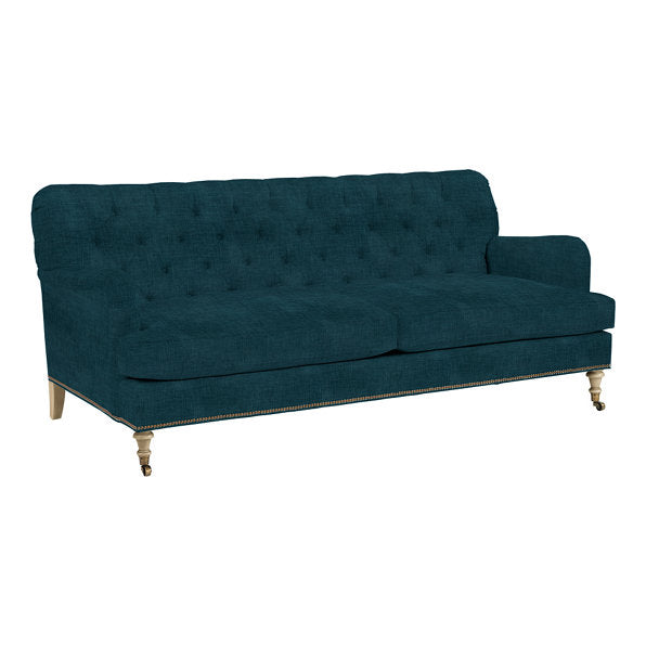 The Maggie Sofa with Brass Nailheads is a dark teal upholstered piece featuring a button-tufted backrest and wooden legs. It boasts two large seat cushions and an elegant design, perfect for enhancing a stylish living room setup.