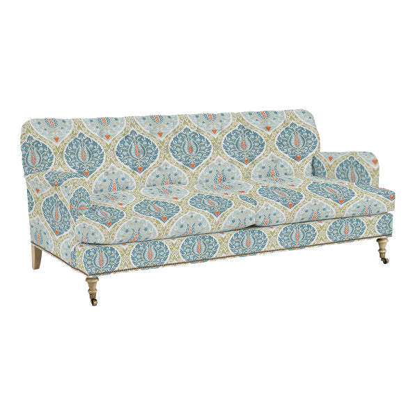 The Maggie Sofa with Brass Nailheads showcases a vintage design adorned with blue and orange paisley motifs on a light beige background. It features wooden legs equipped with small wheels.