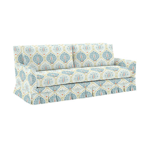 The Suzanne Kasler Mathes Sofa with Box Pleat Skirt is a three-seater featuring a vibrant, patterned fabric design with blue and orange motifs. Its skirted bottom complements the cushions, which are adorned with the same intricate pattern.