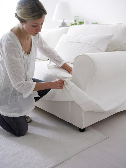 A person adjusts the white slipcover on an UPPLAND Sofa in a bright room. The person, dressed in a white blouse and jeans, is kneeling on a white rug. The UPPLAND Sofa features white pillows, and there's a white lamp visible in the background.