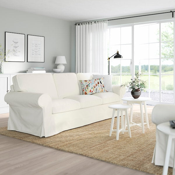 A bright living room featuring the UPPLAND Sofa in white, adorned with a colorful pillow and complemented by white side tables on a beige rug. A large window reveals a lush greenery view outside. The minimalist decor is enhanced with a table lamp, artwork on the wall, and sheer curtains.