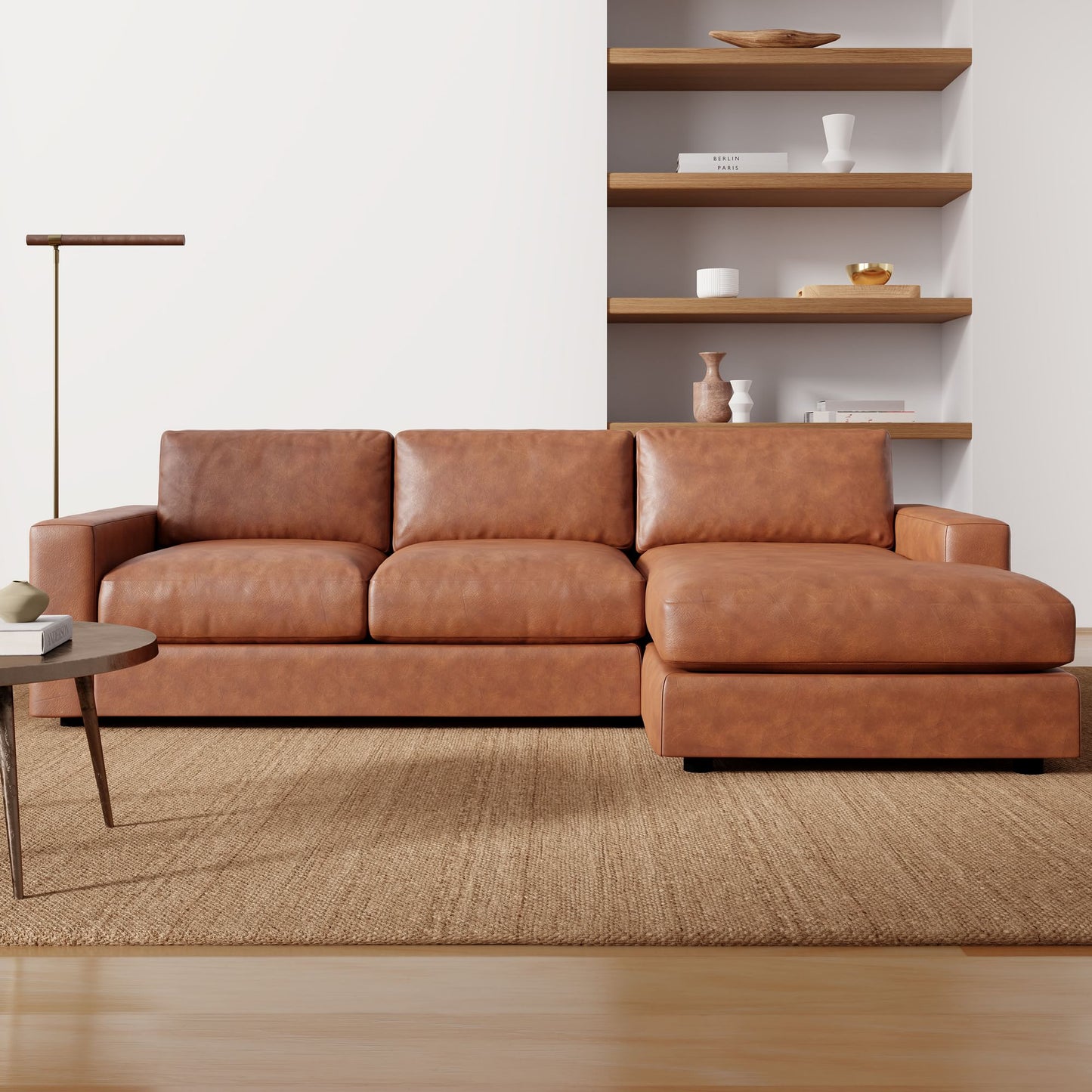 The Urban Leather 2 Piece Chaise Sectional, in a rich brown hue, rests on a textured rug. Behind it, wooden shelves adorned with decorative items accentuate a white wall. A floor lamp and a round coffee table complete the cozy living room scene.