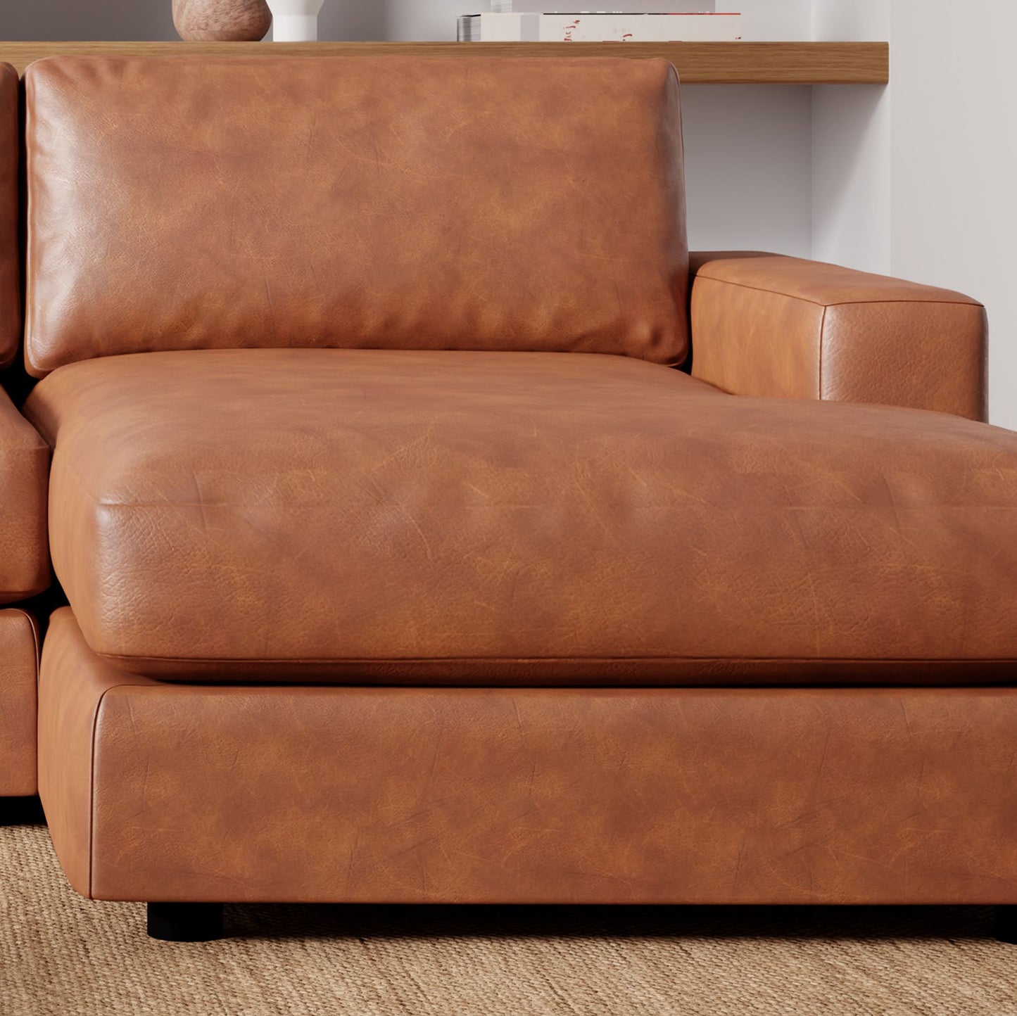 A close-up of the Urban Leather 2 Piece Chaise Sectional in brown, featuring a cushioned armrest. The sectional is placed on a light-colored rug within a living room setup, accompanied by a small wooden shelf showcasing several books and a ceramic vase in the background.