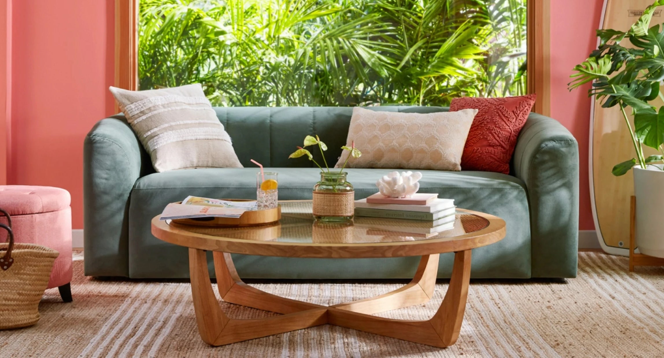 A cozy living room featuring a green sofa with patterned cushions, facing a wooden coffee table with a flower vase, books, and a drink. A pink wall with large windows showcasing lush greenery in the background adds to the vibrant, refreshing atmosphere.