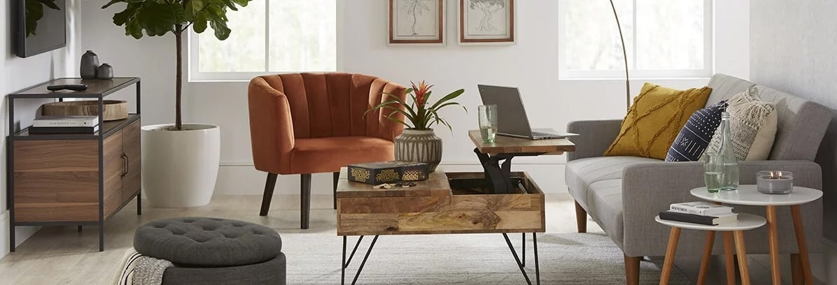 A cozy, modern living room with a gray sofa adorned with yellow and blue cushions, an orange armchair, a wooden coffee table with black hairpin legs, and a white side table. There's a potted plant, framed art, and natural light streaming through the windows.