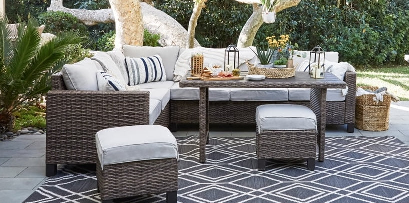 A stylish outdoor patio setup features brown wicker furniture with gray cushions, including a corner sofa, two ottomans, and a rectangular table. The area is enhanced by decorative lanterns, a basket of flowers, a tray of refreshments, and a patterned rug.