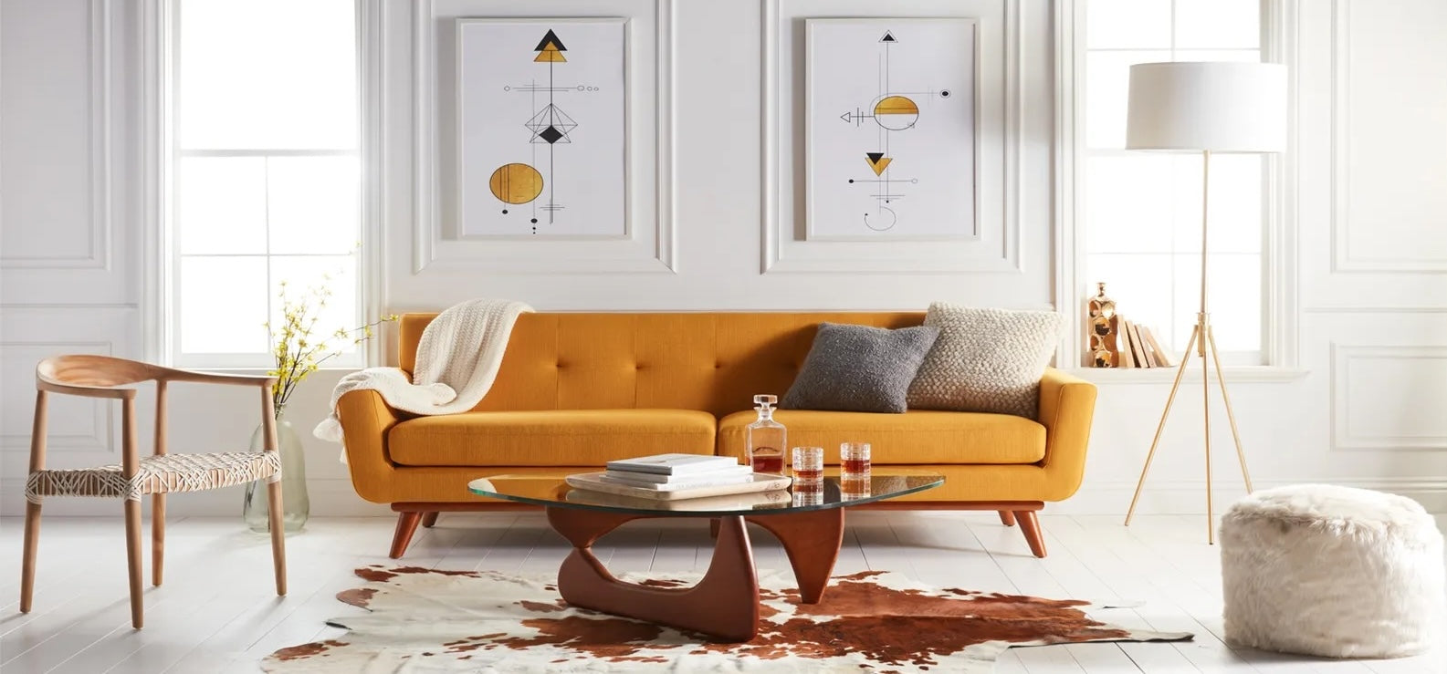 Bright living room with modern decor. A mustard yellow sofa is adorned with neutral pillows and a blanket. Above the sofa are two geometric artworks. The room features a glass coffee table on a cowhide rug, a wooden chair, a tall lamp, and a white pouf.