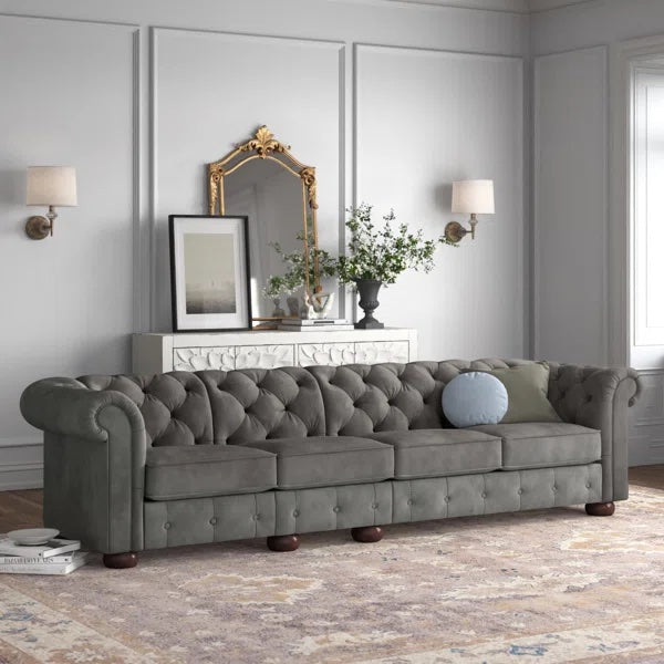 A spacious, elegant living room featuring a long, gray, tufted sofa with rolled arms, accented by a round blue pillow. Behind the sofa is a white console table with framed pictures, a decorative mirror, and a vase with greenery. Two wall sconces provide lighting.