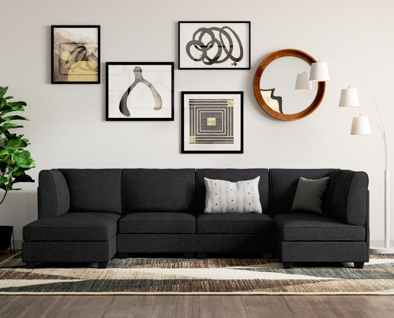 A modern living room features a stylish black sectional couch adorned with two cushions. The wall behind it is decorated with an assortment of framed abstract art pieces. A tall, white floor lamp with three bulbs stands to the right, and a leafy green plant is on the left.