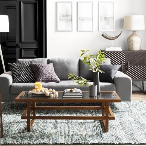 A modern living room features a gray sofa with gray and black pillows, a wooden coffee table with plants, books, and decorative items, and a textured rug. The background includes a black door, framed artwork on the wall, and a sideboard with lamps and decor.