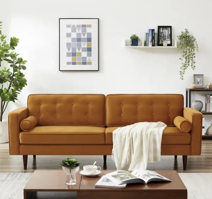 A stylish living room features a mustard yellow sofa with tufted back cushions and bolster pillows. A cream throw blanket drapes over the arm. The room is decorated with plants, a framed abstract artwork, and shelves holding books and decor items. A coffee table displays a book, a teacup, and a small plant.