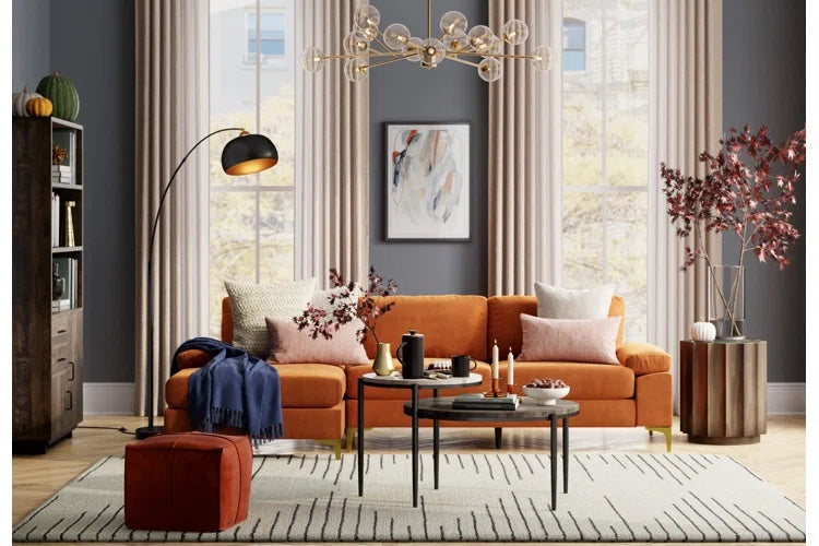 A modern living room features a burnt orange sofa adorned with various pillows and a pink blanket, a black floor lamp, glass bubble chandelier, abstract wall art, two round coffee tables with decor, a striped rug, and large windows with beige curtains.