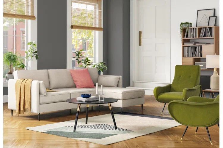 A modern living room with a beige sectional sofa adorned with a pink pillow and yellow throw blanket, a round black coffee table with candles and a bowl, two green armchairs, a white rug, a bookcase, and plants by large windows with natural light filtering in.