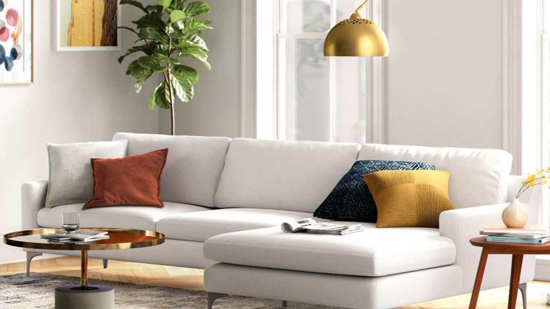 A modern living room with a white sectional sofa adorned with colorful throw pillows in orange, gray, blue, and yellow. A gold floor lamp hangs over the sofa, and a round coffee table with a glass top is in front. A large potted plant and wall art complete the decor.