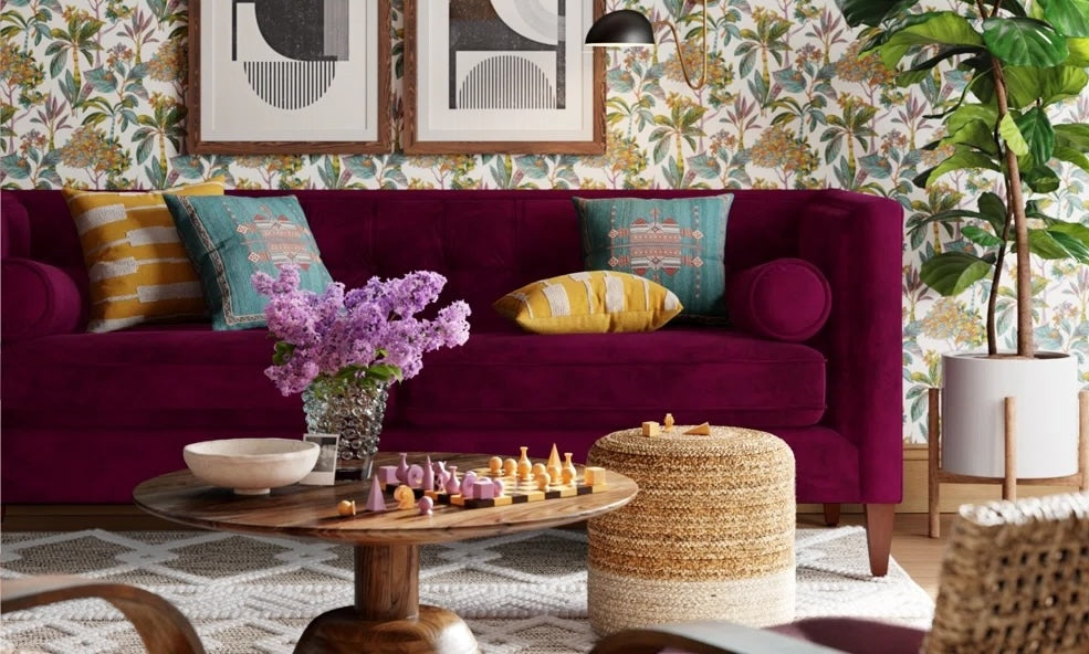 A vibrant living room with a deep purple velvet sofa adorned with colorful cushions, framed abstract artwork on patterned floral wallpaper, a wooden coffee table with a chess set, a potted plant, a woven pouf, and a vase of lilacs on a geometric rug.