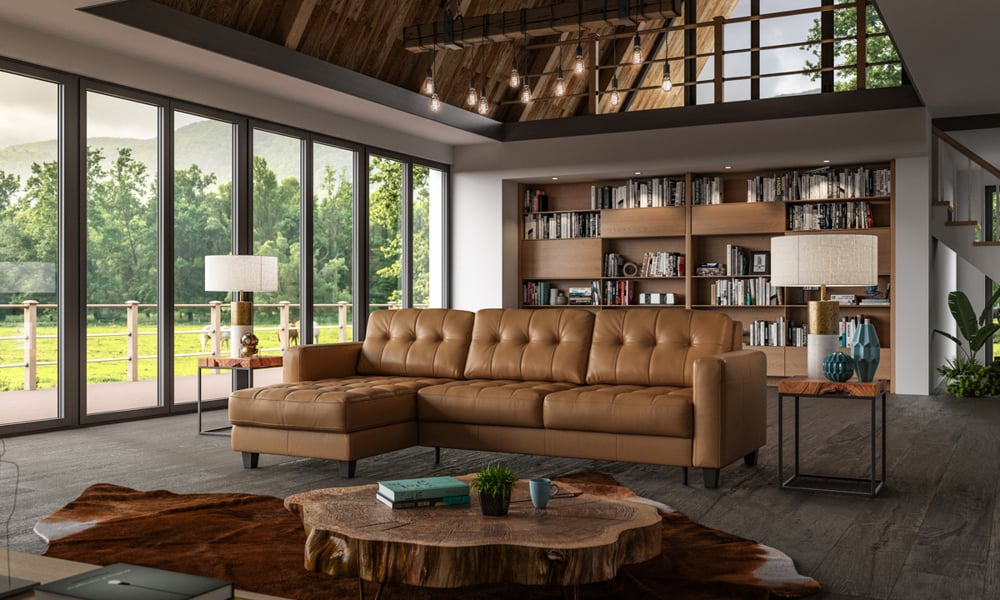 A modern living room features the Luonto Noah Brown Leather Full XL Reversible Sleeper Chaise, complemented by a wooden coffee table, bookshelves brimming with books, and expansive floor-to-ceiling windows that offer views of lush greenery and a wooden deck outside.