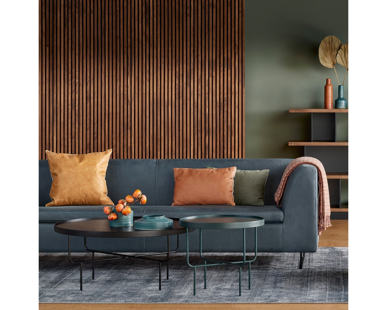 A modern living room showcases a Bonnie and Clyde Sectional Sofa in gray, adorned with brown and green cushions, set against a wooden slat wall. Two round coffee tables display orange vases filled with flowers. A rug spans the floor, while shelves with decorative items add to the background ambiance.