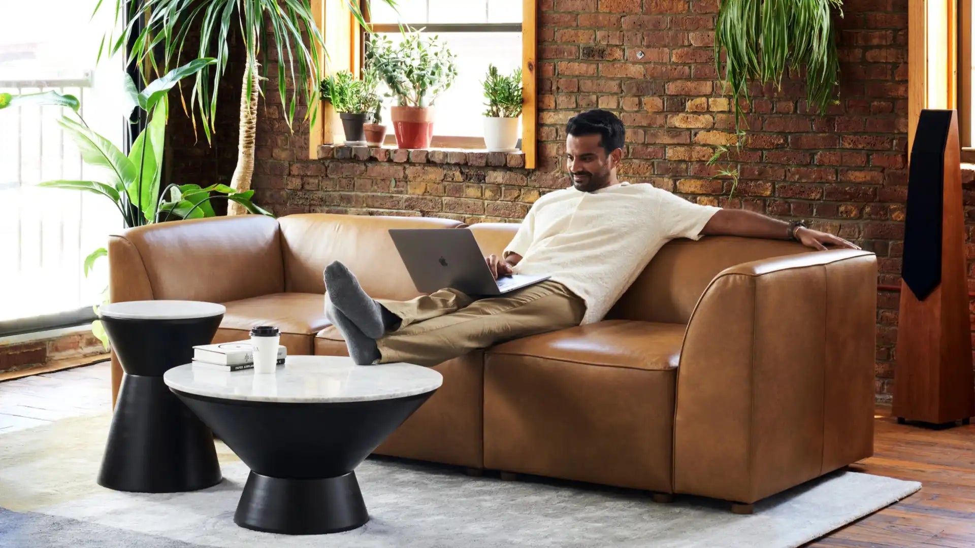 A person relaxes on a Mambo Loveseat, using a laptop. The room is bright with natural light, featuring brick walls and several potted plants. A modern coffee table holds a coffee cup and other items, creating a cozy and inviting atmosphere.