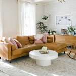 A Beautiful Mess lifestyle and home decor and interior design blog. Modern living room with painted wood panel shiplap wall, sheer white curtains, gold Mid Century space age chandelier, round pedestal coffee table, light brown leather two piece sectional sofa with pink and red multi tone toss pillows.